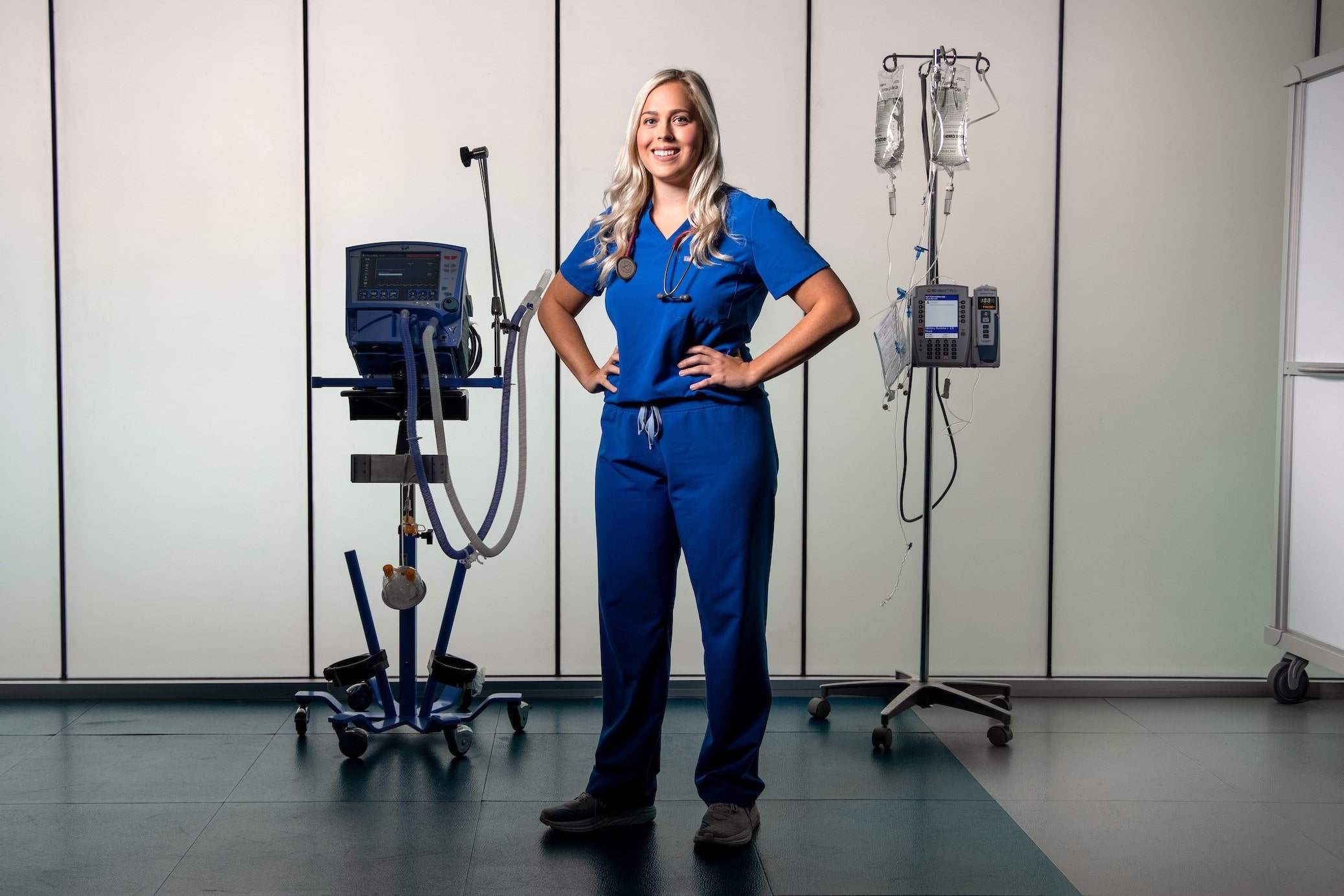 Female CSON Student in blue scrubs with hands on hips standing in front of a medical backdrop surrounded by medical equipment